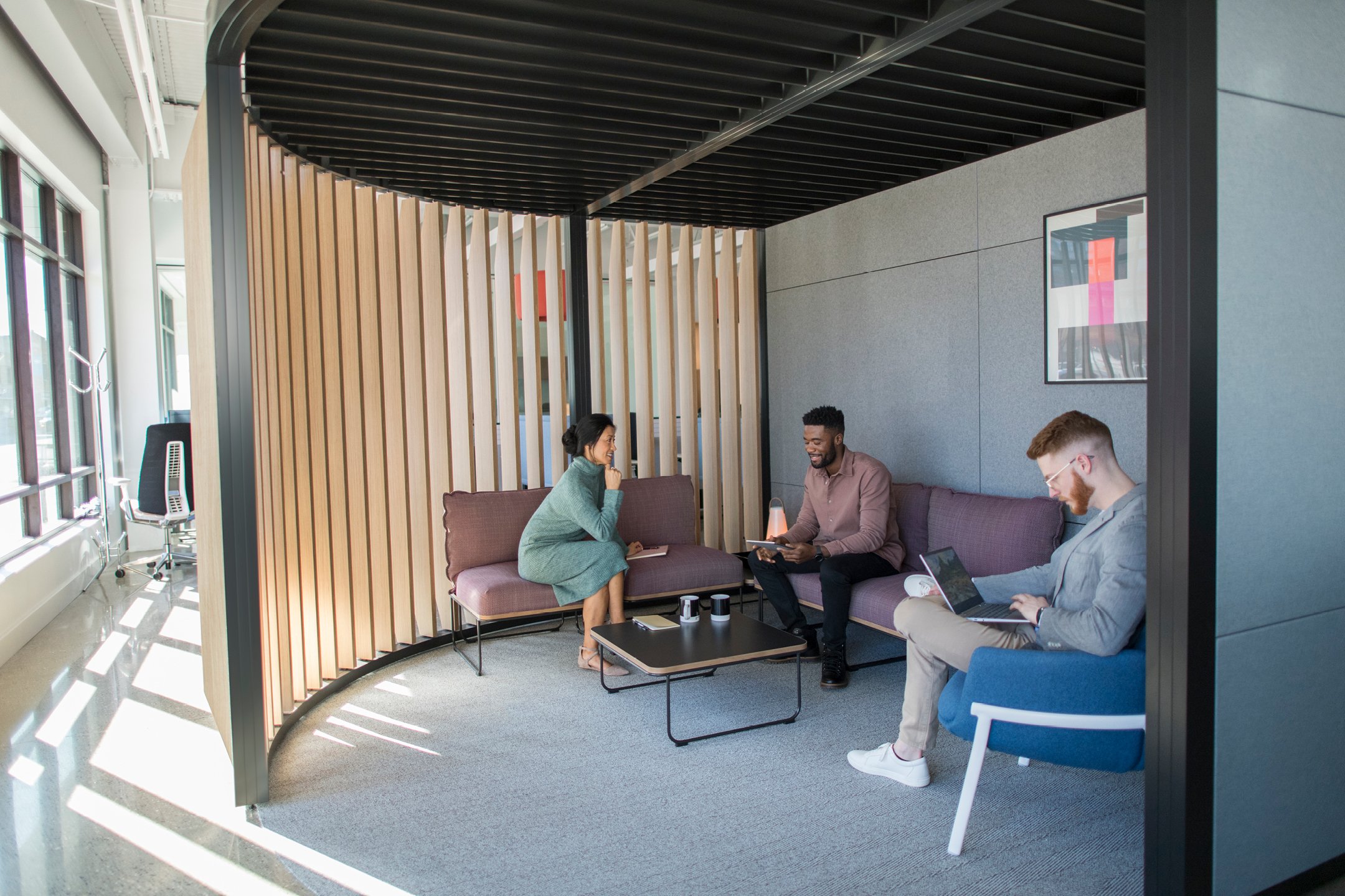 Haworth Pergola Workspace with black trim and grey walls and wood veneer slits for private collaboration room with couch and table and employees working in the office setting